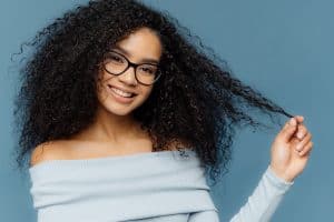 Horizontal shot of lovely smiling Afro American woman holds tips of her curly hair, wears fashionable light blue sweater, transparent glasses, models against blue background. Look at my curls
